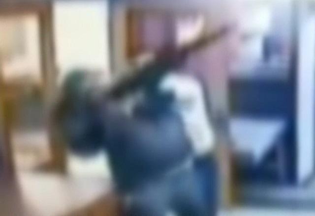 Homem de casaco preto e armado com rifle atirando para o alto e sendo segurado por outro homem em luta corporal