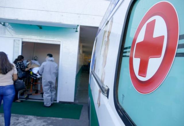 Símbolo de cruz vermelha, estampado em uma Ambulância, na porta de um hospital. No fundo, um homem em uma maca é atendido por profissionais de medicina