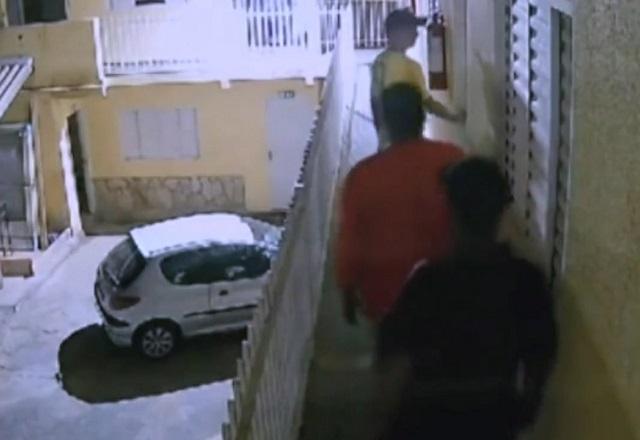 Três homens em fila, em espécie de sacada, na frente da porta de entrada de residência, durante à noite. Ao fundo e no andar térreo, um carro está estacionado